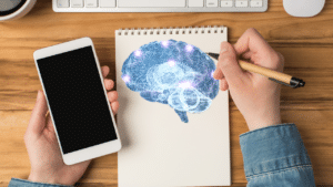 A person is holding a smartphone in their left hand and drawing a brain on a notepad with their right hand.
