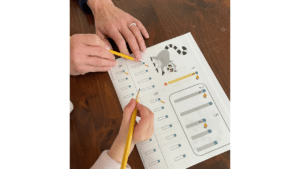 Two pairs of hands holding pencils, working on a worksheet with images and numbered paths, possibly for an educational activity.