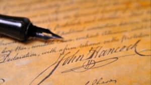 A close-up image of a fountain pen's nib resting on an old parchment with a bold, prominent signature "John Hancock" in cursive handwriting.