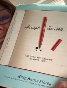 A person holds a book titled "Script & Scribble: The Rise and Fall of Handwriting" by Kitty Burns Florey, with a red pen drawn on the cover, illustrating the nuances of handwriting.