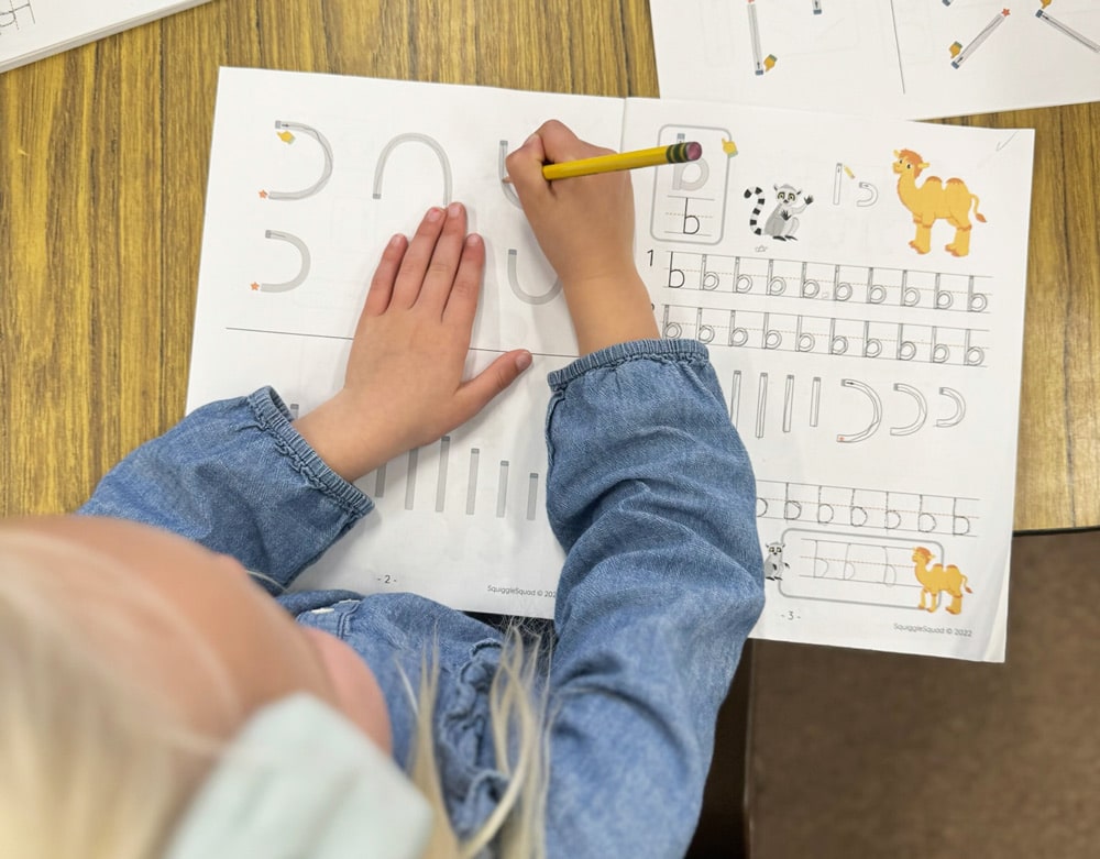 Child practicing writing letters and numbers on a worksheet at a wooden desk.